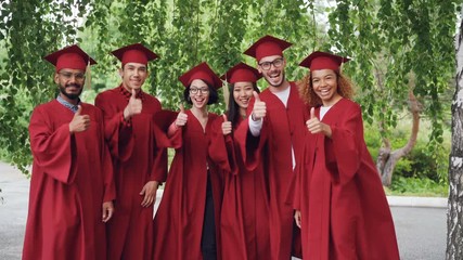 Portrait of excited graduating students multiethnic group standing outdoors in red gowns and mortar-boards and talking then showing thumbs-up and looking at camera.