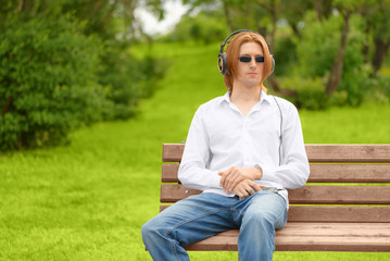 Obraz premium The modern caucasian man is listening to music through headphones on a bench in a park. The melomaniac is wearing a white shirt, the jeans and the sunglasses resting in outdoors.