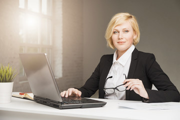 Beautiful blonde adult businesswoman dressed in black stylish suit with eyeglasses working in white wide office on table with laptop