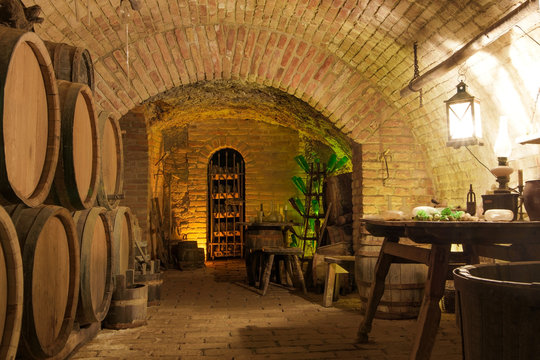 Vintage wine cellar interiors: stack of oak barrels, wooden old-style furniture with glass instruments and rack with dust covered wine bottles