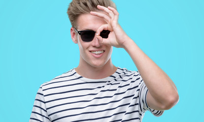 Young handsome blond man wearing sunglasess with happy face smiling doing ok sign with hand on eye looking through fingers