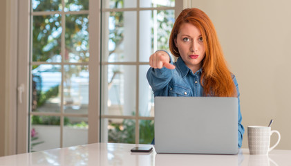 Redhead woman using computer laptop at home pointing with finger to the camera and to you, hand...