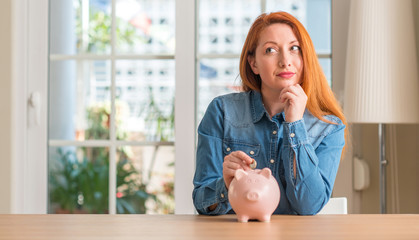 Redhead woman saves money in piggy bank at home serious face thinking about question, very confused...