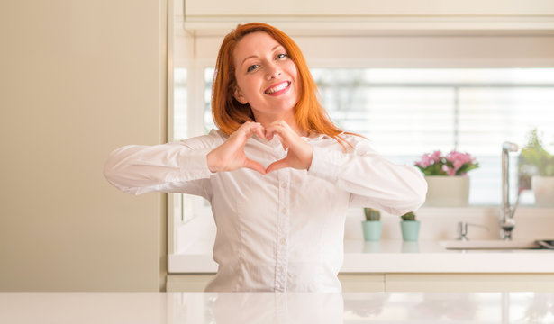 Redhead woman at kitchen smiling in love showing heart symbol and shape with hands. Romantic concept.