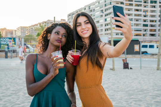 Stylish diverse women with drinks taking selfie on beach