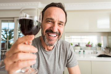 Middle age man drinking a glass of wine with a happy face standing and smiling with a confident...