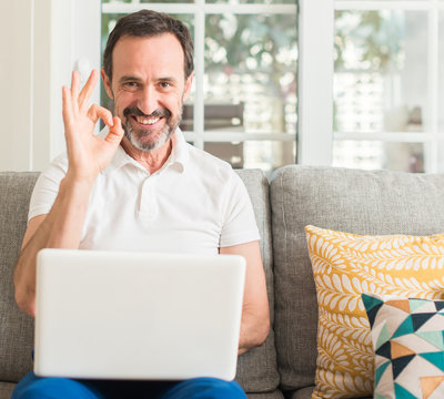 Middle age man using laptop at sofa doing ok sign with fingers, excellent symbol