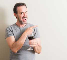 Senior man drinking red wine in a glass very happy pointing with hand and finger to the side