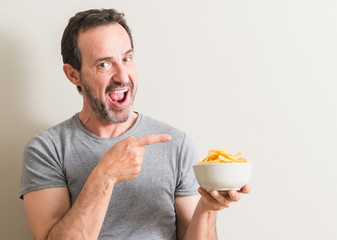 Senior man eating potato chips very happy pointing with hand and finger to the side