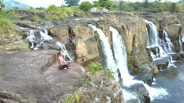 girl sits in pose lotus on high steep cliff by waterfall