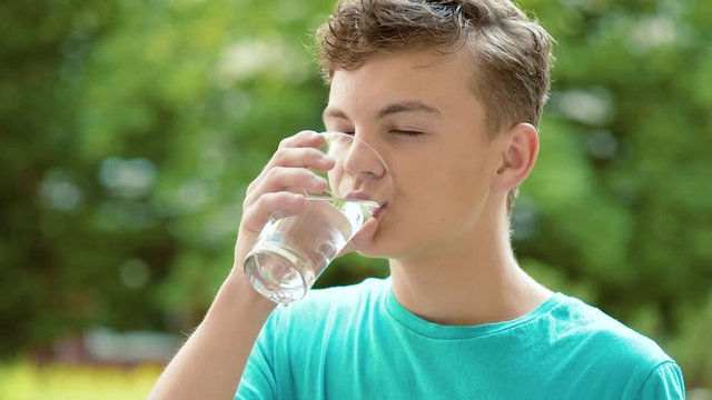 Happy teen boy drinking water from glass in summer park. Cute teenager drinks cold water - outdoors portrait. 