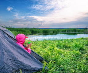 Girl in pink jacket sits at the exit of tent with mug and looks 