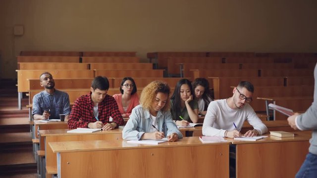 Professor is reading lecture to group of multiracial students standing in classroom with papers and speaking while young people are listening to him and writing.