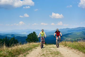 Young smiling bikers man and woman in professional sportswear riding bikes on dusty mountain road under bright blue sky on magnificent mountain range background. Tourism and active lifestyle concept.