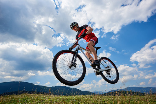 Athletic sportsman cyclist in professional sportswear and helmet flying in air on his bicycle on summer blue sky with white clouds and distant mountains on background. Outdoor extreme sport concept