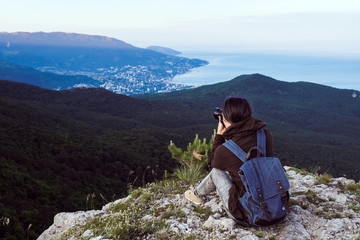 Naklejka premium A traveler girl sits on top of a high mountain, takes pictures at the camera and looks at the sunset.