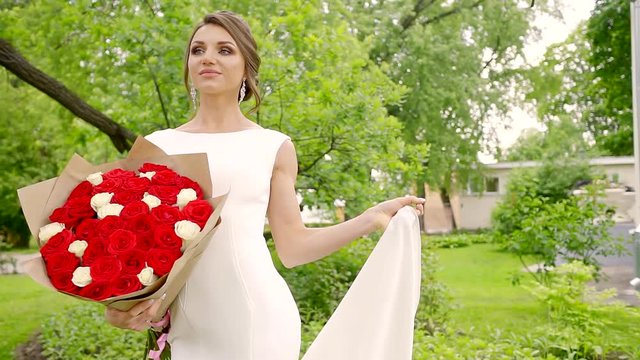 a young bride stands on the nature in a wedding dress, in her hand have bouquets of roses