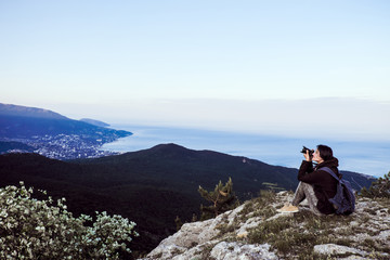 Naklejka premium A traveler girl sits on top of a high mountain, takes pictures at the camera and looks at the sunset.
