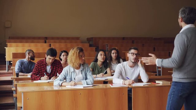 Tutor is talking to students sitting at desks in spacious college lecture hall, young people in casual clothes are asking questions. Education and university concept.