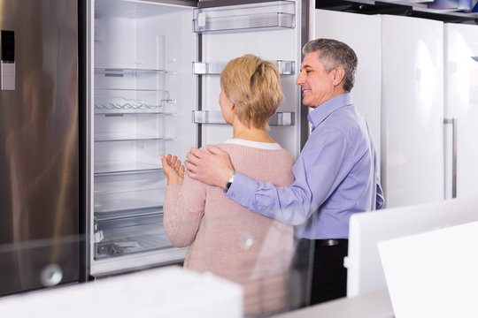 interested mature married couple in shop of household appliances are choosing home modern refrigerator