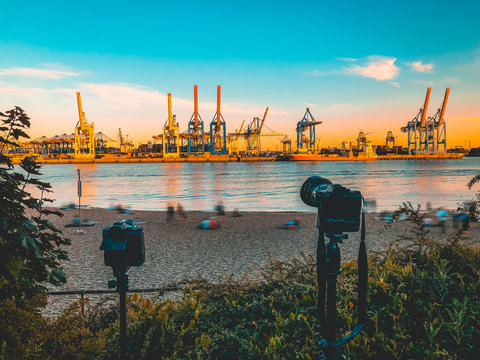 two cameras taking pictures of industrial port at hamburg with beach in the foreground and blurred tourists