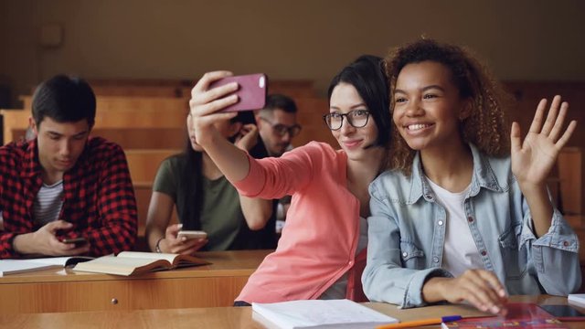 Cheerful girls students are taking selfie with smartphone sitting at tables at college, women are posing making hand gestures and hugging. Friends and technology concept.