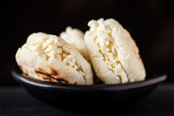 Colombian traditional white corn arepa stuffed with grated cheese in a black ceramic dish on black background