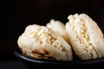 Colombian traditional white corn arepa stuffed with grated cheese in a black ceramic dish on black background