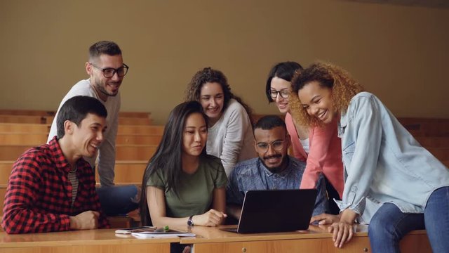 Cheerful men and women are working with laptop looking and pointing at screen, talking and laughing sitting at desks in university lecture hall. Students and technology concept.