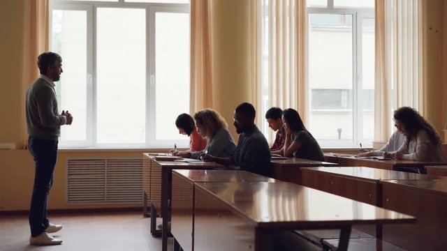 Bearded man university teacher is reading lecture to adult students and gesturing , young people are writing and listening to him. Higher education and youth concept.