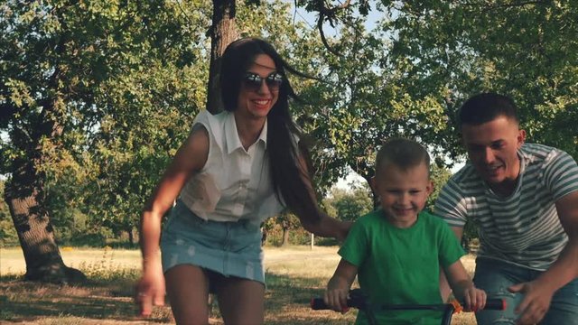 Happy family, mother, father and son are walking by nature, the child is riding a bicycle, his parents help him to accelerate. Happy parents and the baby are smiling. The video.
