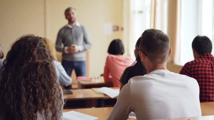 Participant of youth conference young man in glasses is raising hand and talking to tutor while other people are looking at educator and listening to conversation. - Powered by Adobe