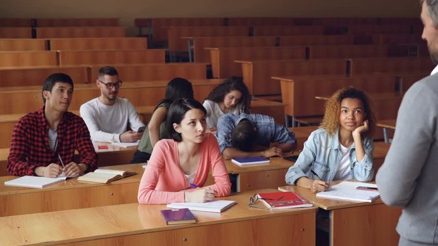 Tired mixed race student is sleeping during lecture at college, his classmates are waking him up, he is standing and talking to teacher, young people are laughing.