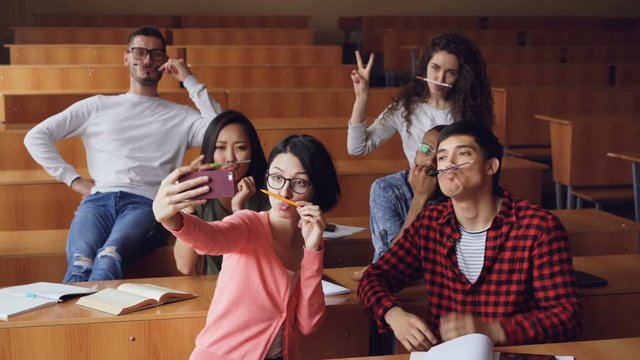 Playful young people are taking funny selfe using pens and pencils as moustache, posing and showing hand gestures thumbs-up and v-sign sitting in classroom.
