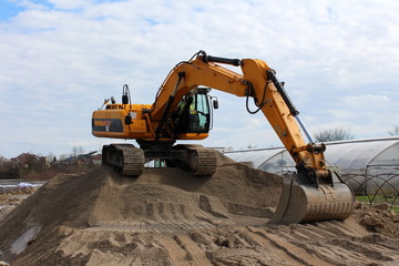 Excavator standing on top of sand pile waiting for truck with greenhouse, houses, river and cloudy blue sky in background
