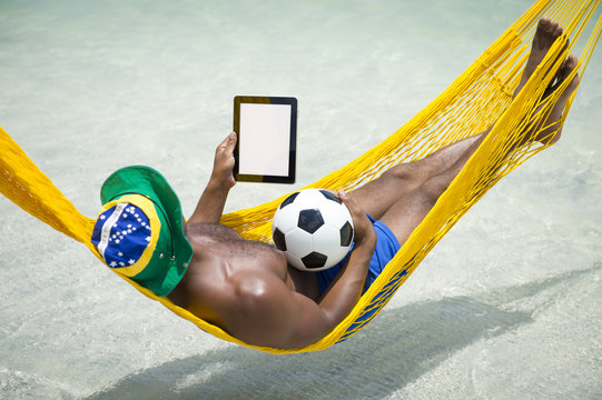 Brazilian footballer wearing flag hat and using tablet computer with a soccer ball relaxing in bright sunlit hammock on beach over the sea.
