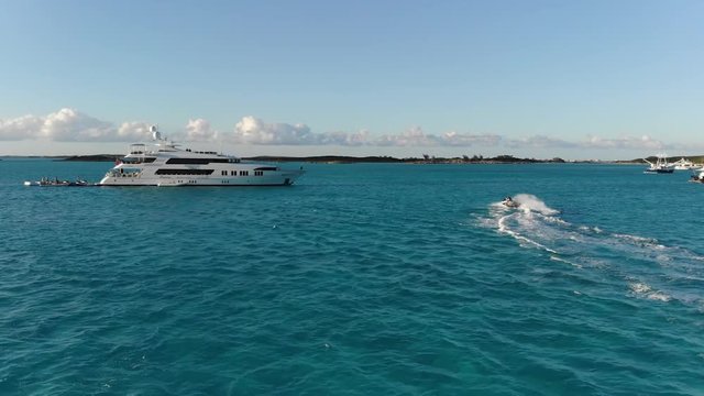Mavic Air drone follows couple through blue Caribbean Sea in front of large yacht and fisherman boats. Aerial view of Exumas, Bahamas water sports.