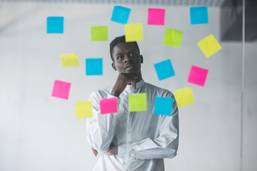 Young afro american business man standing in front of stickers glass wall and looking on futures...