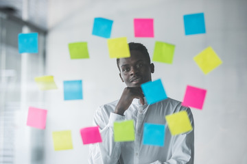 Young afro american business man standing in front of stickers glass wall and looking how to achive goals at his office place