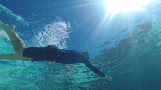 Diver swims over underwater camera in half wetsuit as sun peaks through the ocean water. GoPro Hero Six mounted on the sea floor.