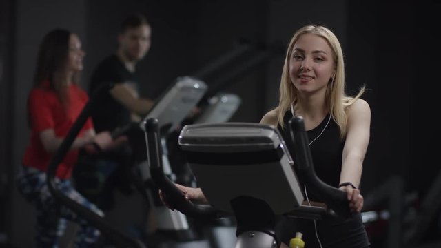 cute girl in blak sport wear vigorously works on exercise bike and approve showing thumbs-up in the new gym against the background of engaged people