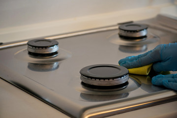 A human hand in a blue rubber glove washes the gas stove with a detergent.