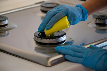 A human hand in a blue rubber glove washes the gas stove with a detergent.
