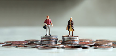 A miniature man and a miniature woman standing on top of a pile of coins of the same height.