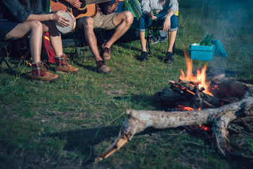A group of Asian friends sitting on chairs, singing, playing a guitar and drinking some beer and water together outside the tent near the fire while they has camping on Weekend holiday.