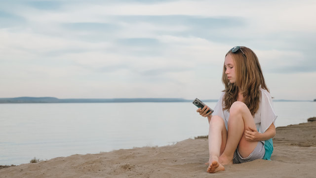 One beautiful teenage girl with brown hair outside on a beautiful summer day. The woman is sitting on the beach. The girl rewrites in her smartphone.