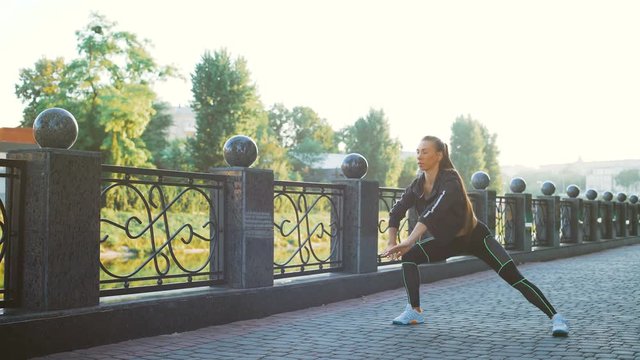Active woman exercising and stretching in city park. Attractive fit girl training warming up at morning outdoors. Healthy, fitness, wellness lifestyle. Sport, cardio, workout concept