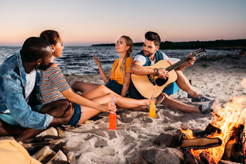 happy young multiethnic friends enjoying guitar and spending time together on sandy beach at sunset