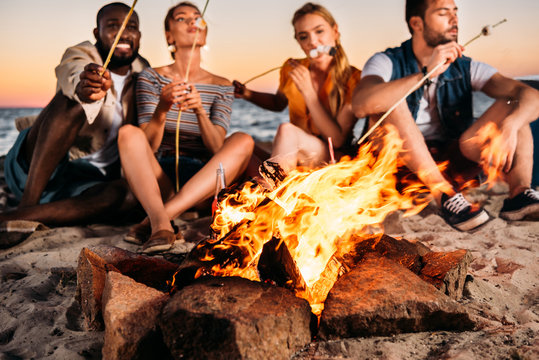 young friends roasting marshmallows at bonfire while sitting at sandy beach at sunset - Powered by Adobe