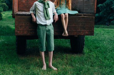 a barefooted girl in a green dress sits on the back of a car and hugs a barefoot, next to a standing guy, a summer day in the village. teenagers in the village. rural life of adolescents